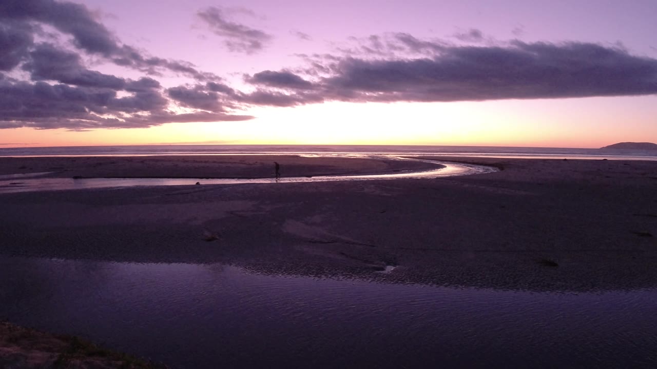 A wide Pismo Beach sunset frame shows the channel bending through dark sand toward the bright horizon, with the small person visible near the middle distance.