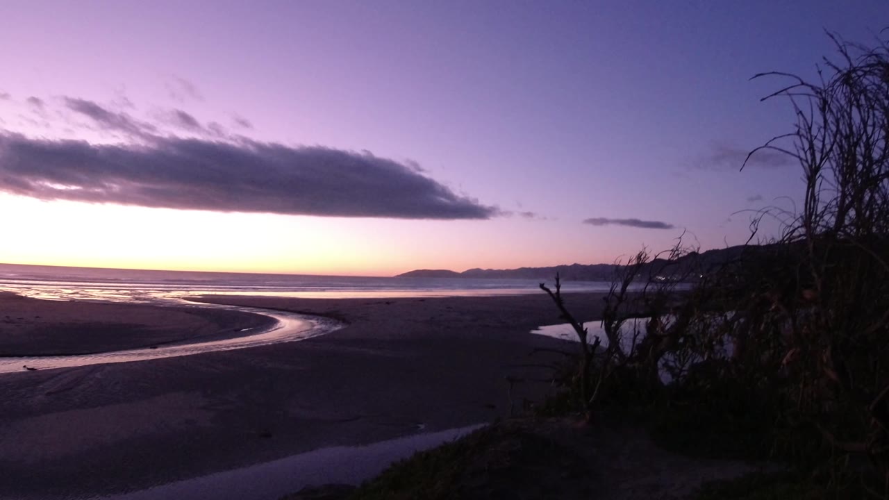 Wide Pismo Beach dusk frame with the river channel bright against dark sand, the Pacific horizon beyond it, a long cloud overhead, and no visible people.
