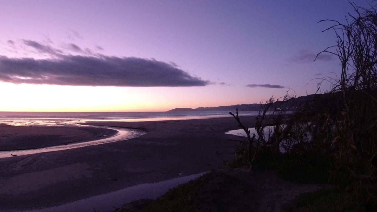Pismo Beach shoreline at sunset with the river mouth sweeping through the midground, dark brush filling the right edge, low hills on the horizon, and no visible people.