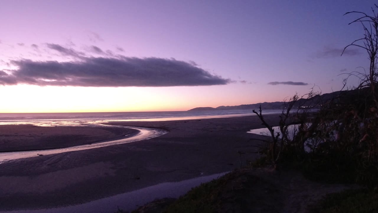 Pismo Beach at dusk with broad dark sand, a smooth river channel curving toward the ocean, pastel light along the horizon, sparse right-edge brush, and no visible people.