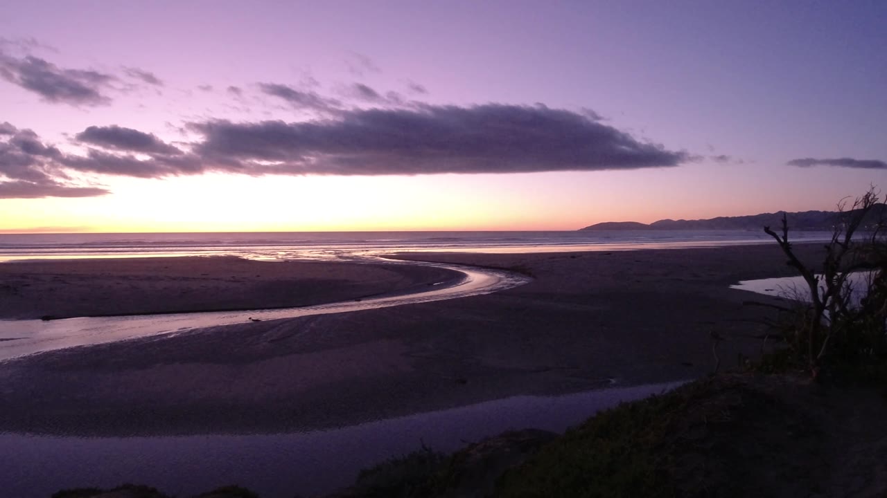 Elevated twilight view over Pismo Beach, California, showing the beach flats, a bright reflective channel, layered ocean water, distant hills, dark cloud cover, and no visible people.