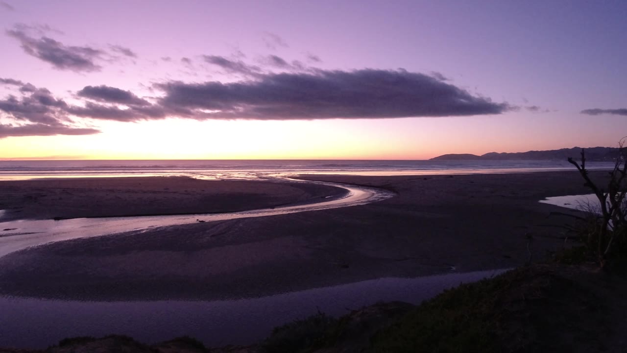 Pismo Beach, California, seen from above or a high viewpoint, with the water channel bending through the sand flats, dark foreground vegetation at right, glowing horizon light, and no visible people.
