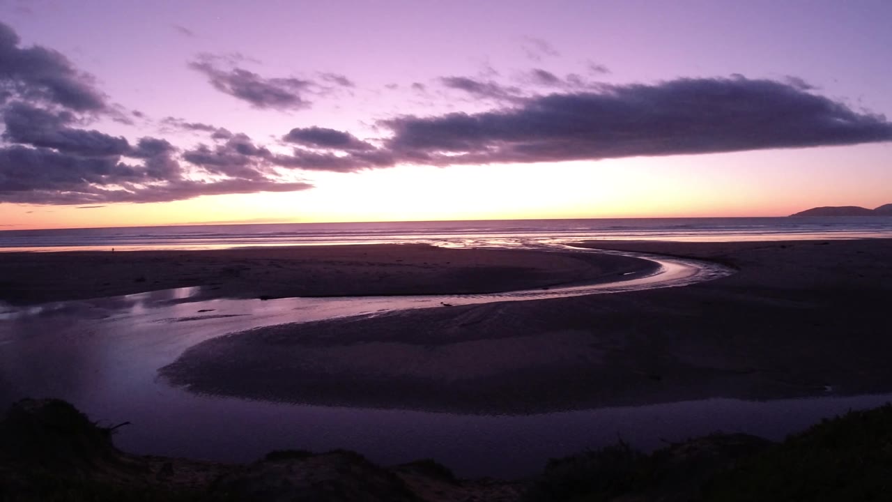 Pismo Beach, California coastal flats under a purple sky, with the tidal channel bending across the frame, a bright sunset band, distant land, and no visible people.