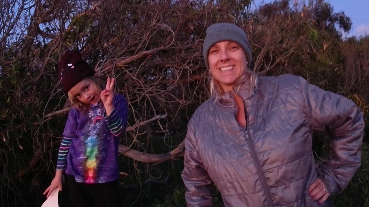 In Pismo Beach, a blonde woman in a gray beanie smiles beside a young girl making a peace sign in front of dense bushes.