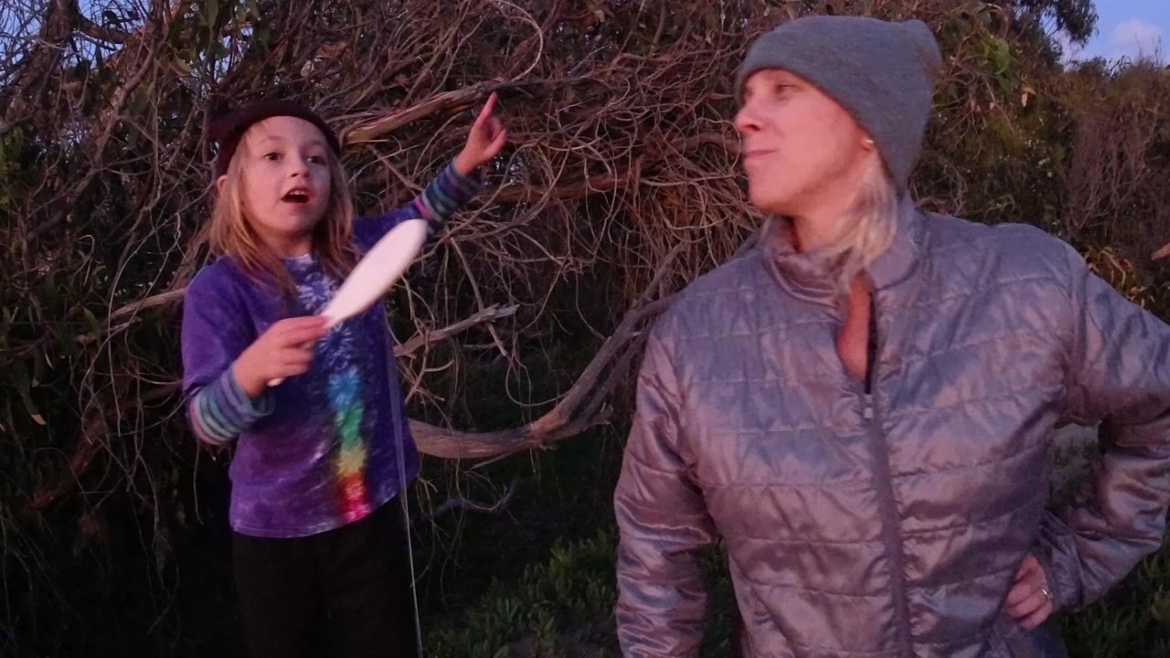 At 0:00 in Pismo Beach, a child in a purple shirt raises one hand while holding a white object beside an adult woman in a gray beanie near dense branches.