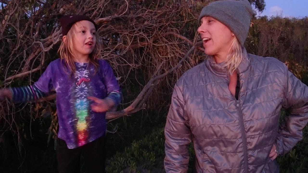 At 0:02 in Pismo Beach, the child moves closer toward the adult woman, both with open expressions, against a background of tangled branches and green vegetation.