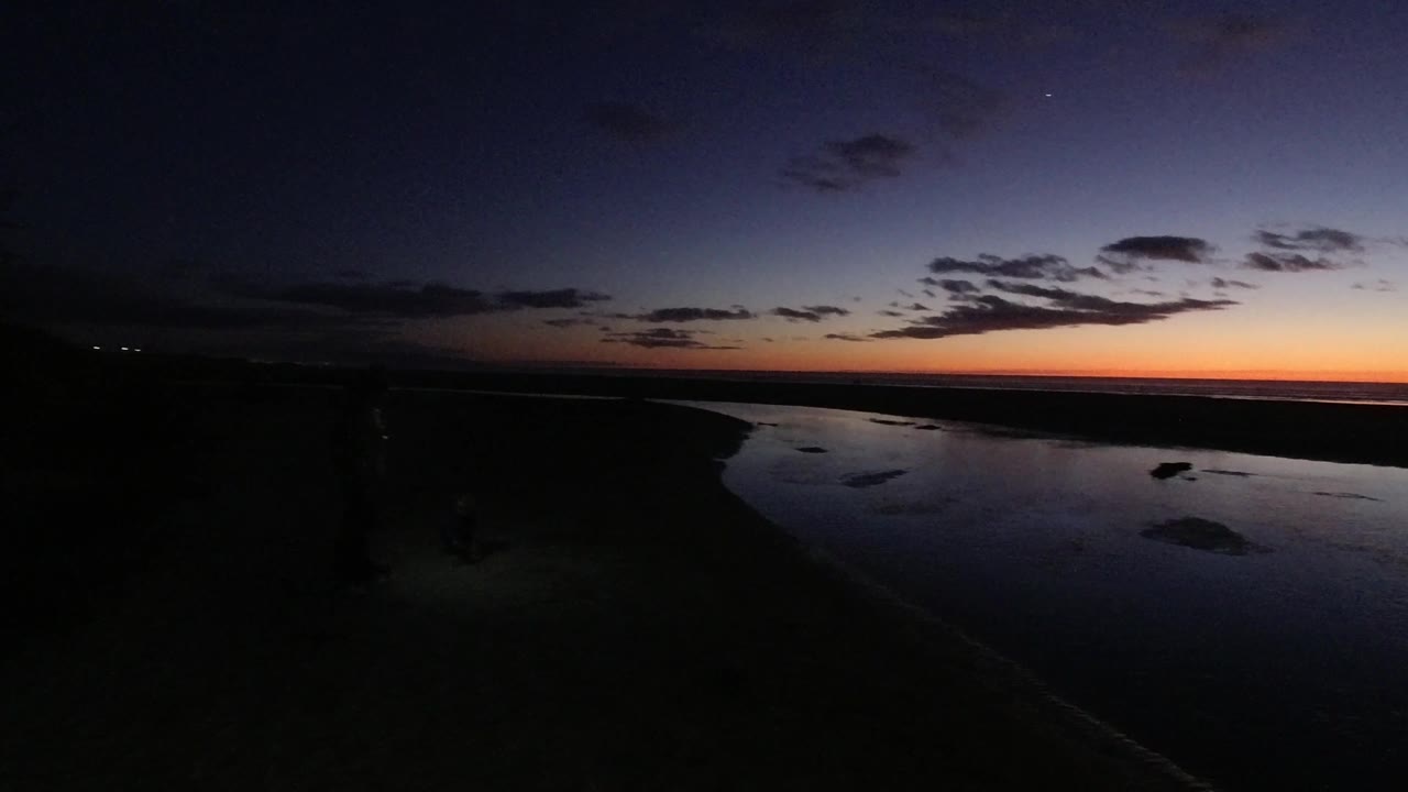 At 0:02, the Pismo Beach shoreline remains dark while reflective water and the orange horizon glow sit beneath a cloud-dotted evening sky, with no visible people.
