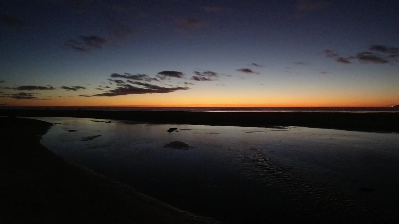 Pismo Beach, California sunset frame showing a broad dark water surface, a flat shoreline silhouette, a narrow orange glow, and no visible people.