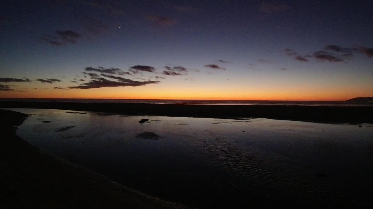Pismo Beach, California at 0:03, with a brighter strip of orange along the horizon, dark tidal water below, scattered clouds above, and no visible people.