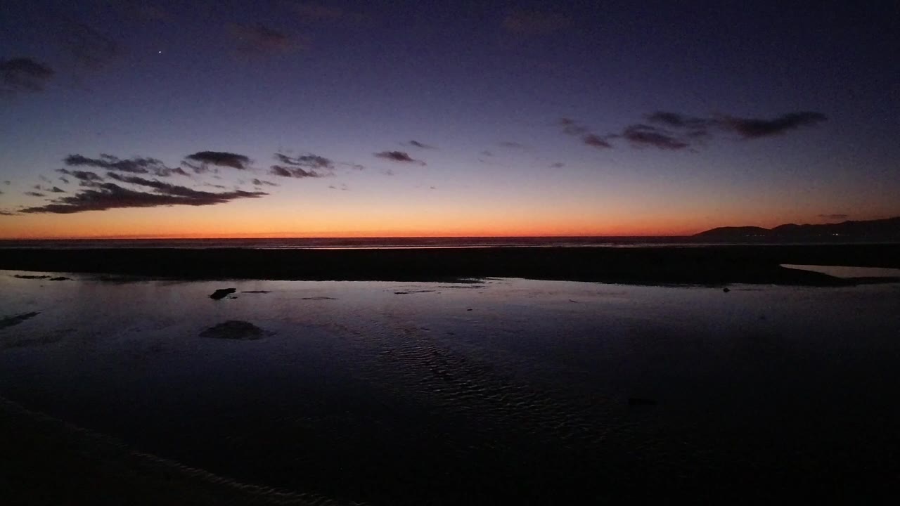 Pismo Beach sunset frame with a thin orange horizon, dark reflective beach water, scattered clouds, distant hills, and no visible people.