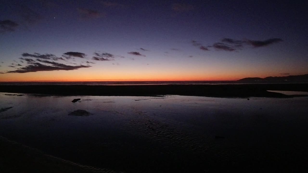 Pismo Beach at dusk with a narrow orange band across the horizon, darker clouds overhead, reflective foreground water, and no visible people.