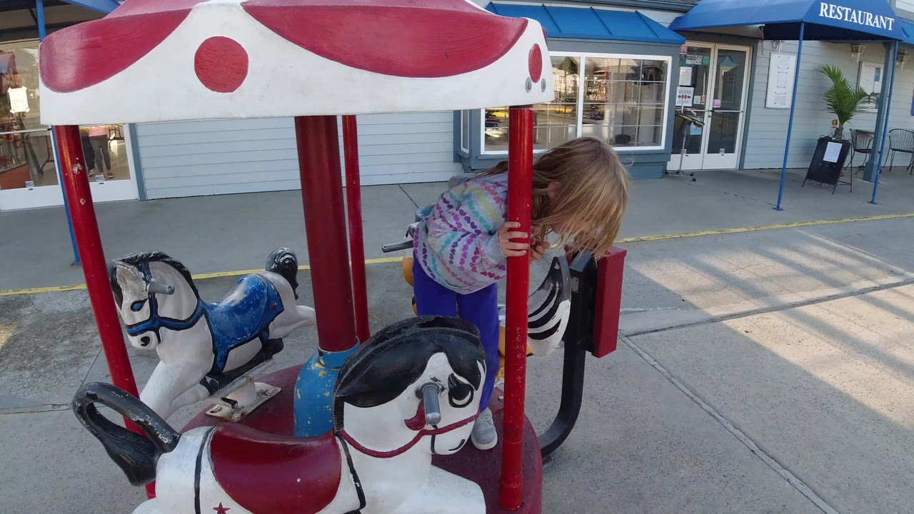 A young girl in a striped sweater leans near the red center pole of a small carousel horse ride in Pismo Beach, with blue-trimmed storefront windows behind her.