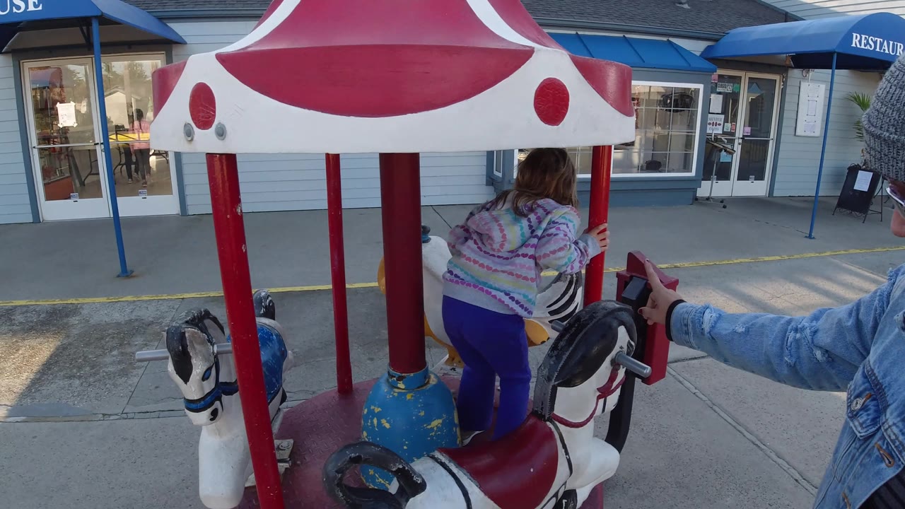 The girl climbs onto a white carousel horse as an adult arm reaches toward the ride controls, with the storefront and blue awnings in the background in Pismo Beach.