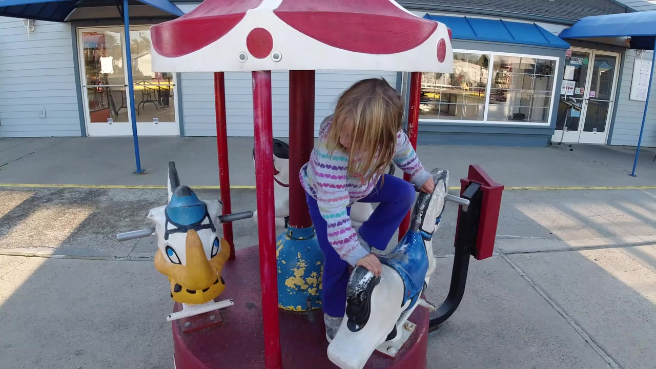 A young girl in a striped sweater climbs onto a blue and white carousel horse in Pismo Beach, California, with a red canopy pole and storefront windows behind her.