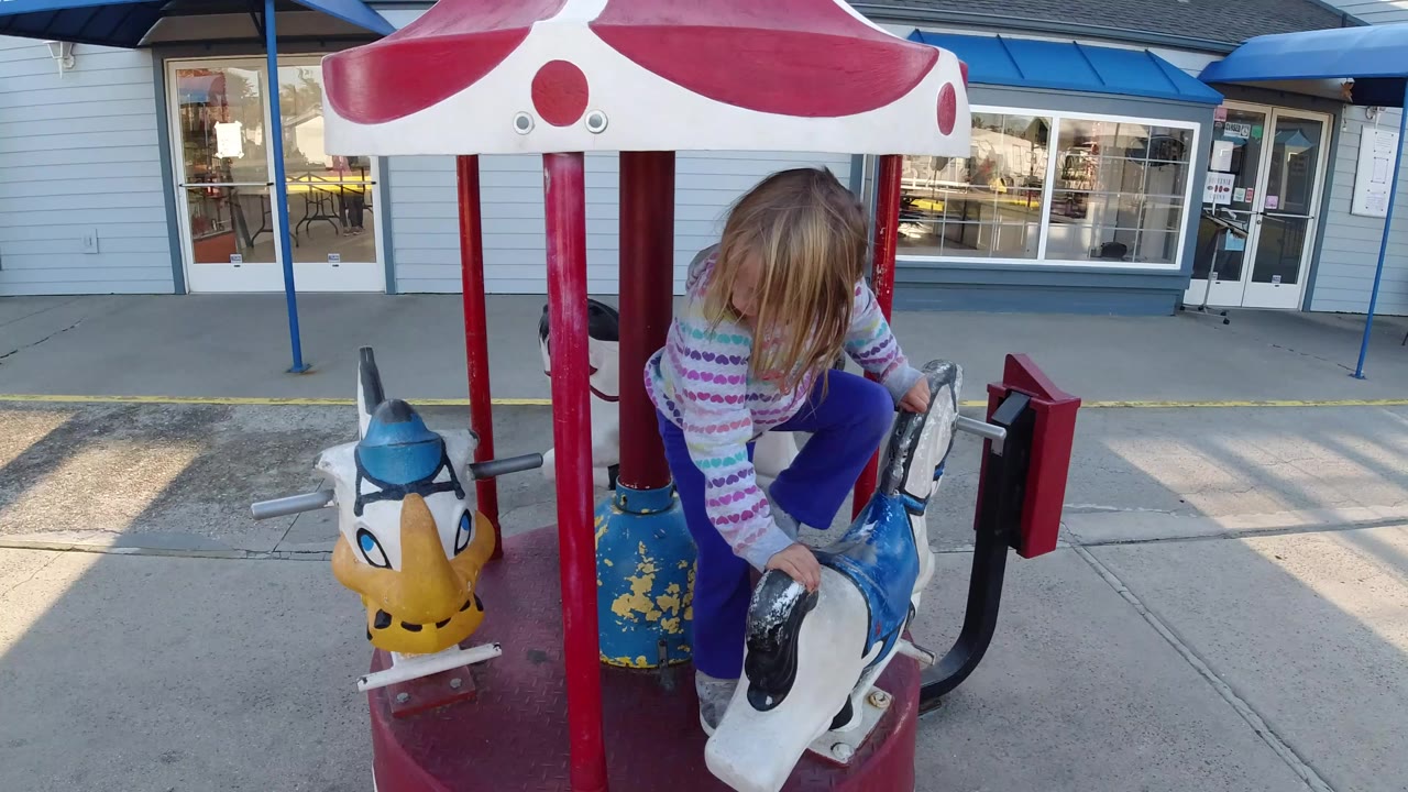 The girl sits on a white carousel horse in Pismo Beach, California, framed by red poles with the storefront window line and blue trim in the background.