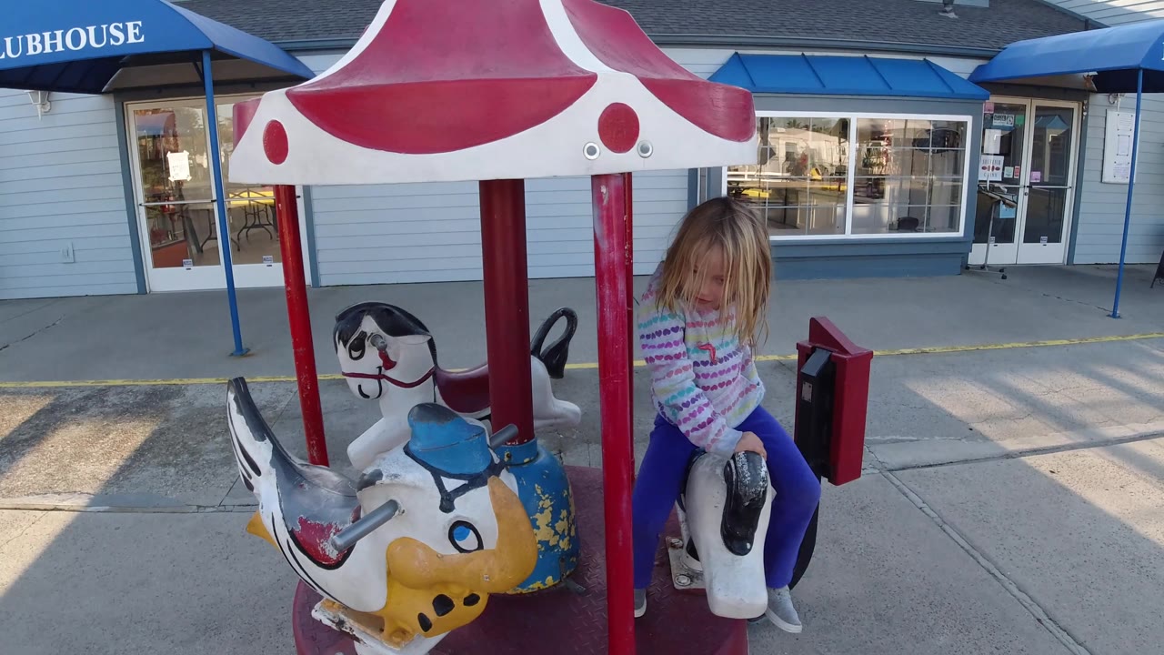 A young girl rides beside a yellow carousel animal under the red and white canopy in Pismo Beach, California, with the blue CLUBHOUSE awning visible above the walkway.
