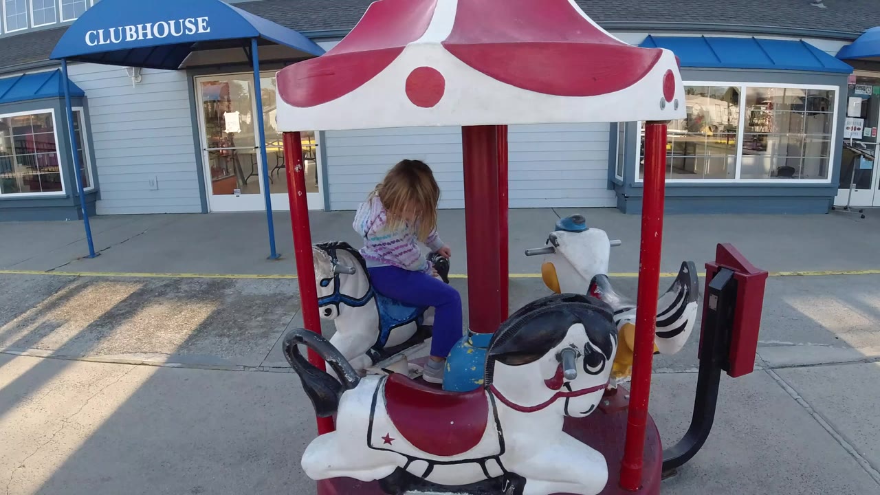 A child in a striped hooded sweatshirt sits on a white carousel animal at a small outdoor ride in Pismo Beach, California, with a CLUBHOUSE awning behind the ride.