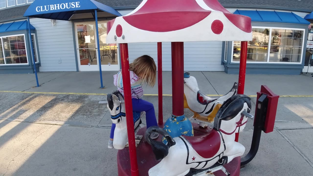 The child is partly turned away on the small carousel in Pismo Beach, California, with multiple painted animal seats, red support poles, and long shadows across the pavement.