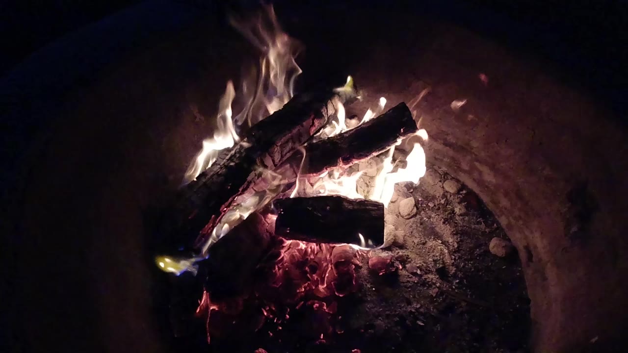 At 0:00, burning firewood glows inside a dark fire pit in Pismo Beach, California, with white flames rising from charred logs and no visible people.