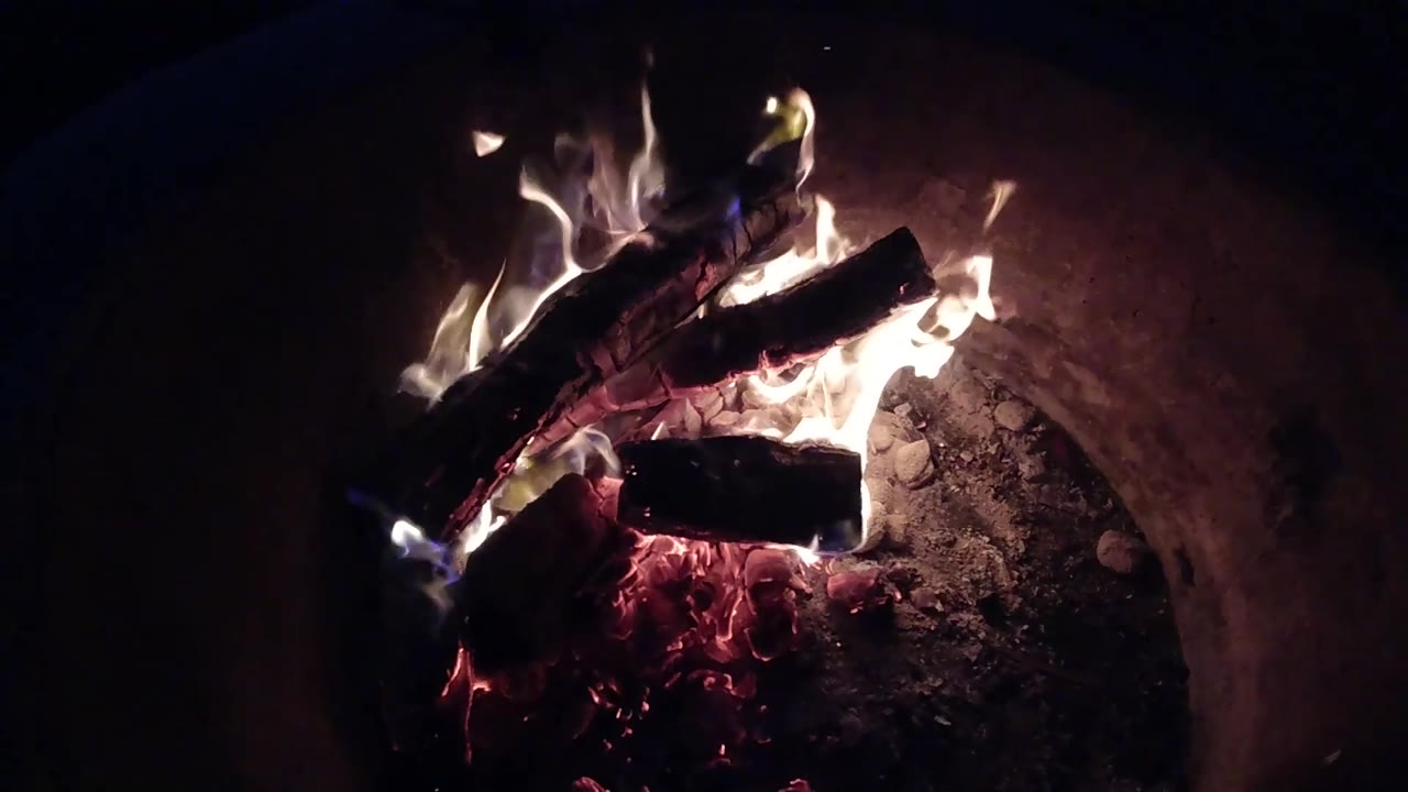 At 0:02, bright flames lean across charred firewood inside the circular fire pit in Pismo Beach, California, with no visible people.