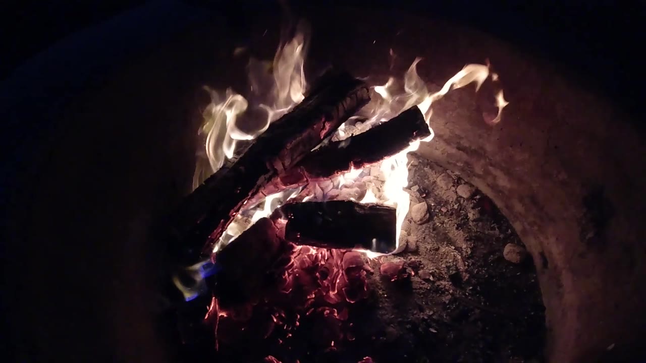 At 0:03, pale flames rise from the top log while a lower blackened log sits over red embers in the Pismo Beach fire pit, with no visible people.