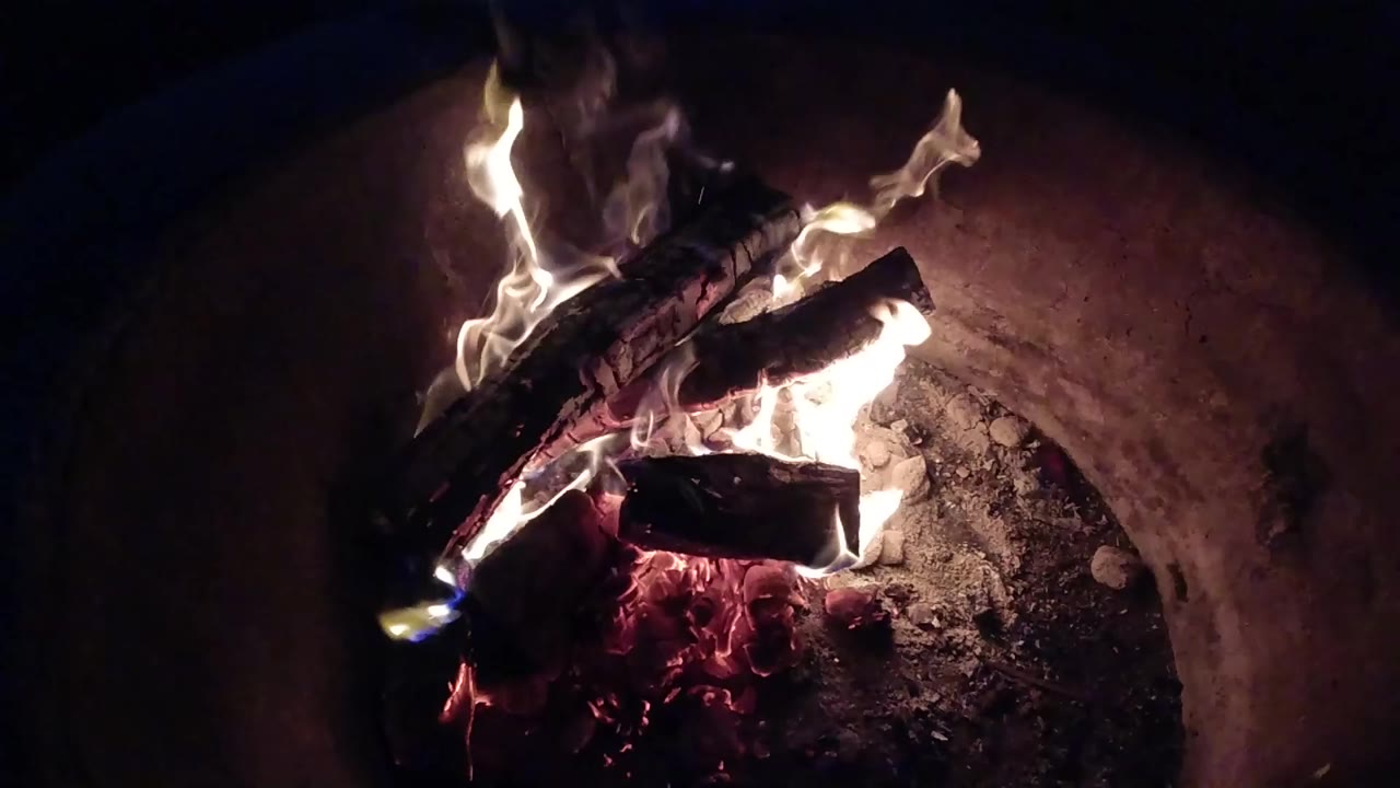 Burning logs rest over red embers inside a round fire pit in Pismo Beach, with pale flames rising at the start of the clip and no visible people.