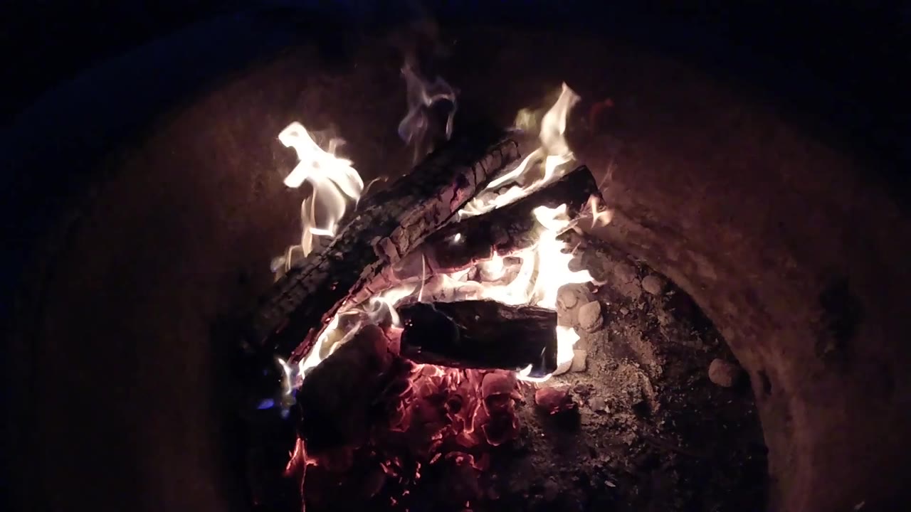 Bright white flames flare along the right side of the stacked logs in the Pismo Beach fire pit, with the ash bed visible behind them and no visible people.