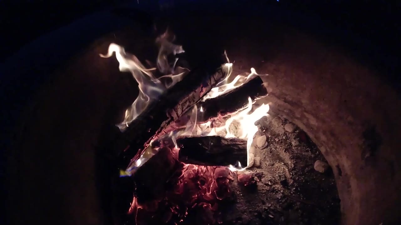 A taller flame rises on the left of the charred log stack in the Pismo Beach fire pit, with red embers concentrated underneath and no visible people.
