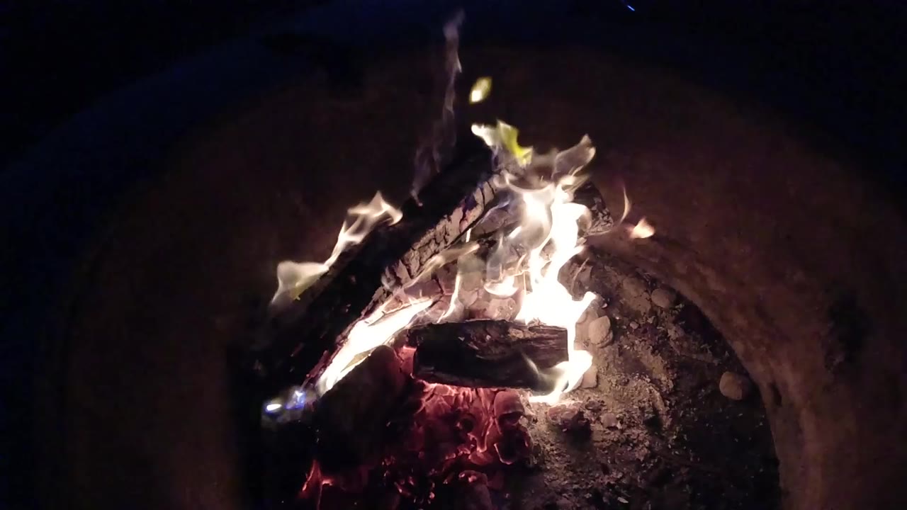 At 0:00, bright flames rise from crossed logs in a rounded fire basin in Pismo Beach, California, with red embers below and no visible people.