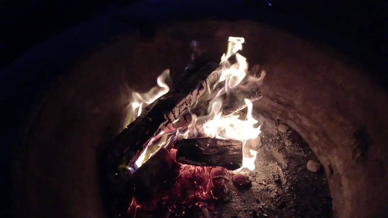 At 0:02, white flames curl over charred logs near the top of the basin, with a reddish ember bed below in Pismo Beach, California, and no visible people.