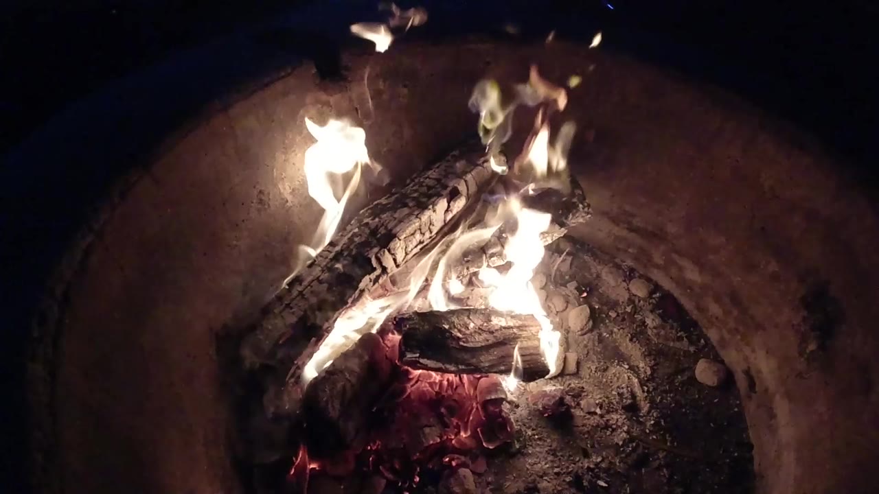 At 0:00, burning logs rest over red embers inside a round fire pit in Pismo Beach, California, with no visible people.