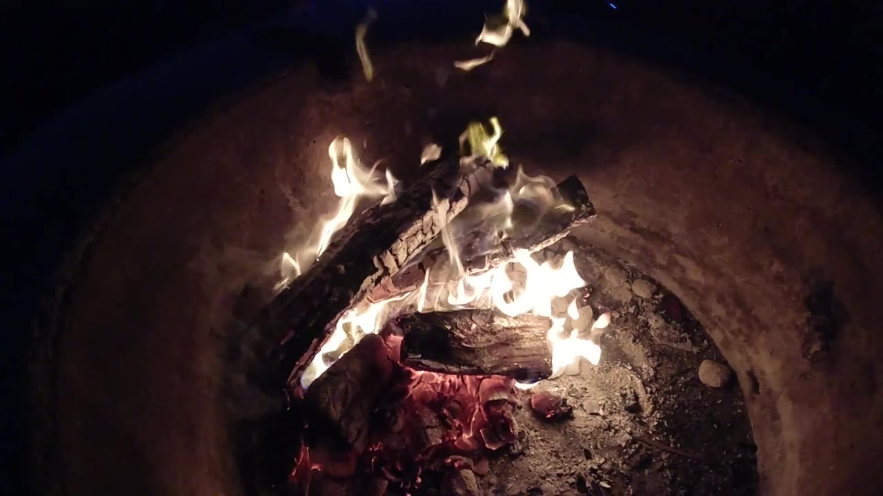 At 0:02, bright flames spread across charred logs above a bed of ash and red coals in the Pismo Beach bonfire, with no visible people.