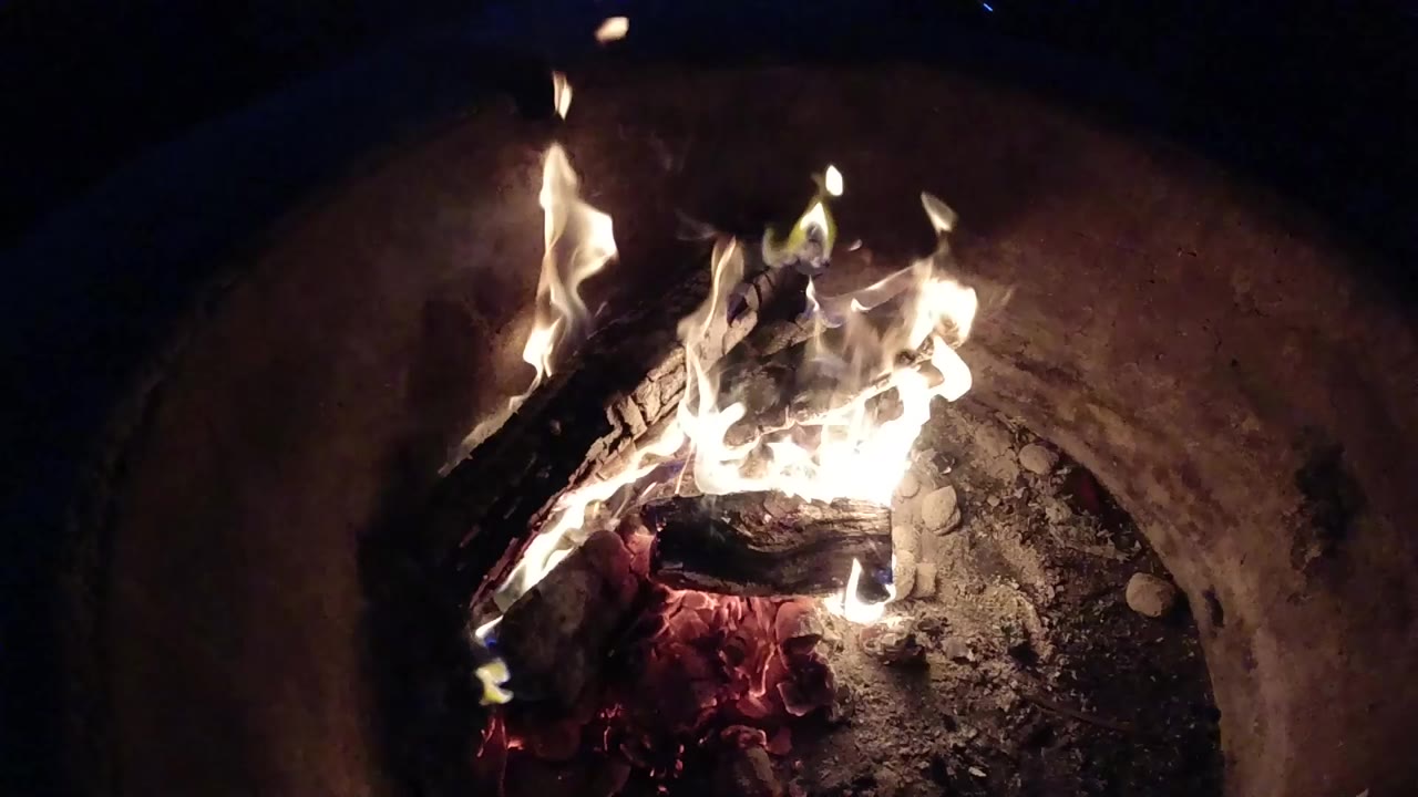 At 0:01, flames climb from the center of charred logs while embers glow below inside the Pismo Beach fire pit, with no visible people.