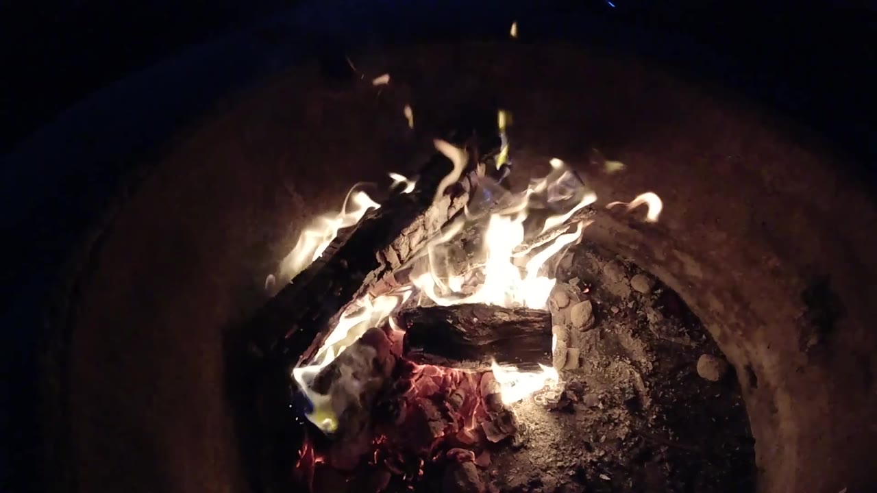 At 0:02, bright flames spread across stacked wood in the dark grey pit, with ash and small stones visible and no visible people.