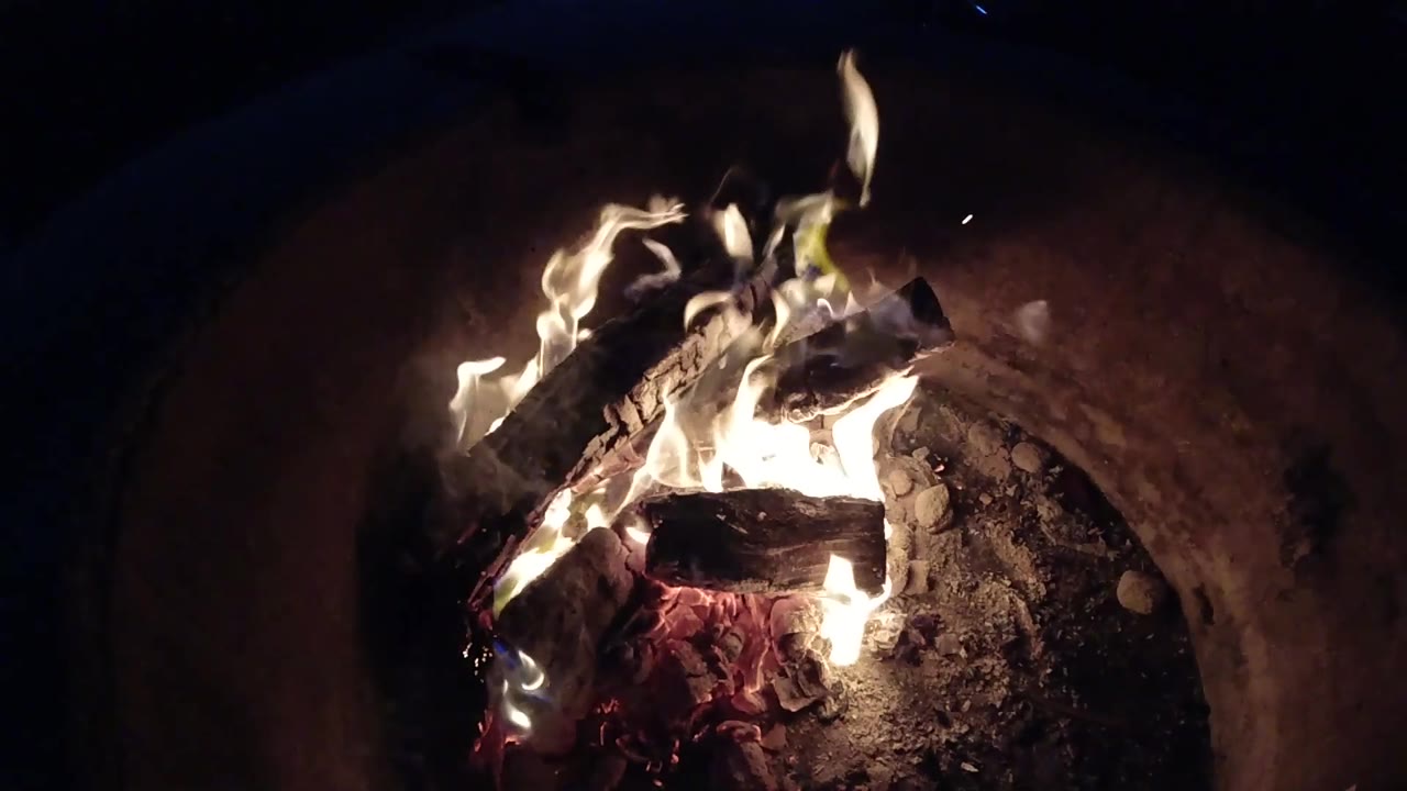 At 0:03, a taller flame curls above the burning wood while the red ember bed glows at the lower edge of the pit, with no visible people.