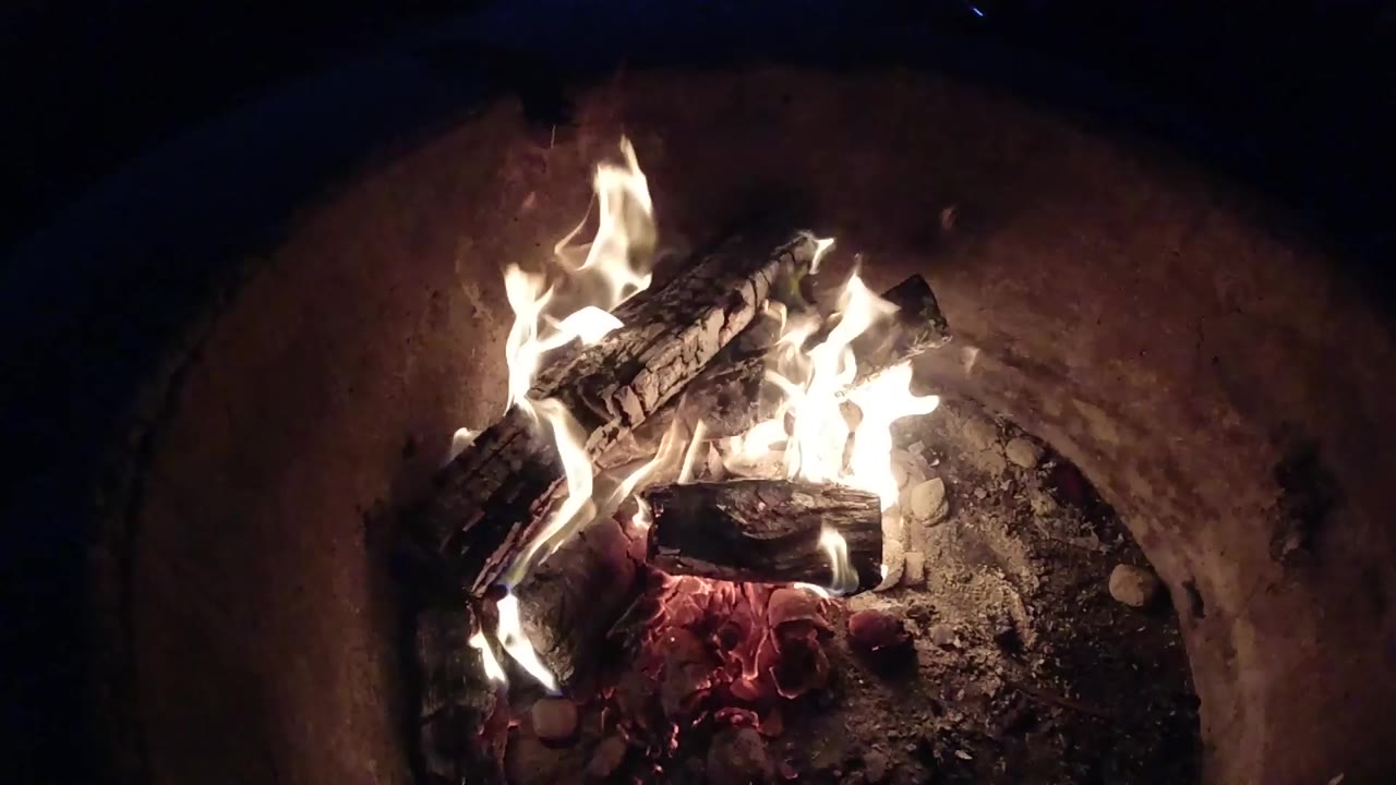 At 0:01, tall white flames wrap around charred logs over red coals in the Pismo Beach, California footage, with no visible people.