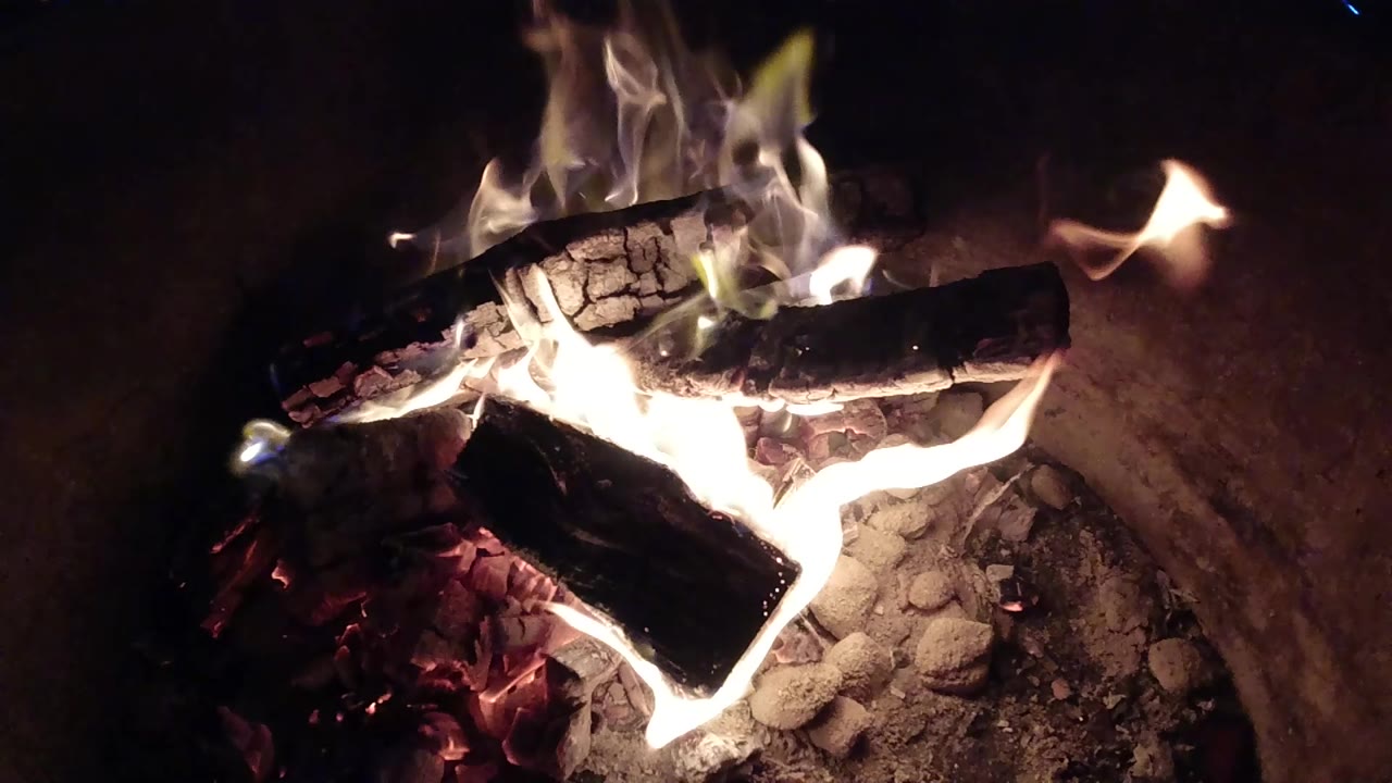 At 0:01, flames curl around two blackened logs inside the barrel in Pismo Beach, California, with a dark upper background and no visible people.