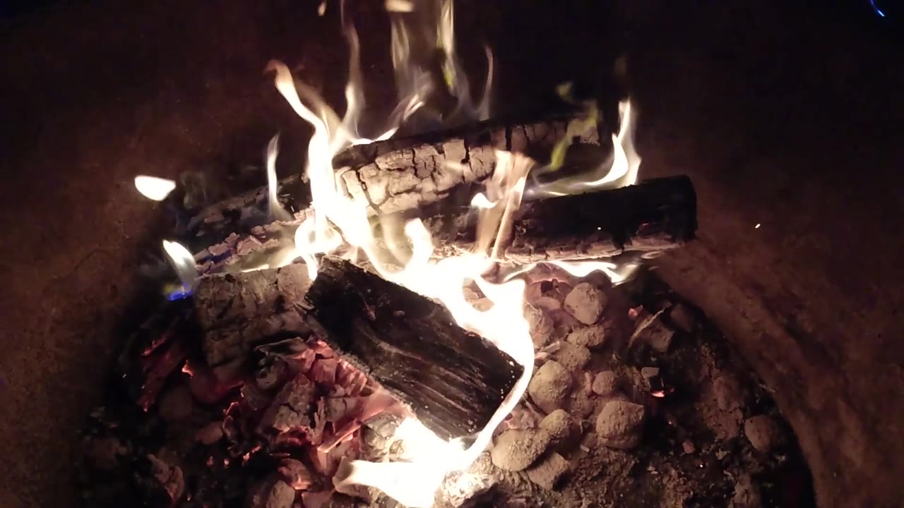At 0:03, charred wood burns over pale ash and red coals inside the barrel in Pismo Beach, California, while the right side falls into darkness and no people are visible.