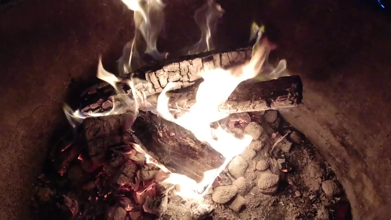 Tight high-angle view of split logs burning over ash and red embers in a Pismo Beach fire pit, with no visible people.