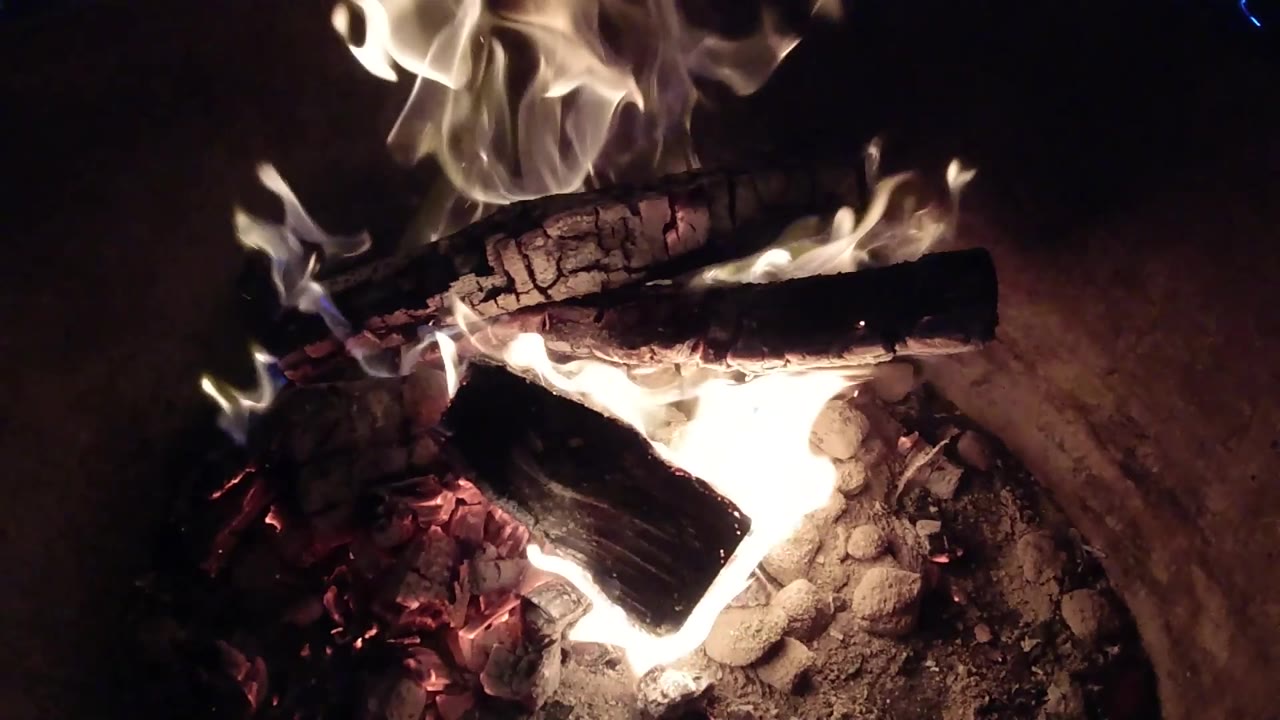 At 0:02, a stronger flame flare appears beside the stacked logs in the Pismo Beach fire pit, with dark surroundings and no visible people.