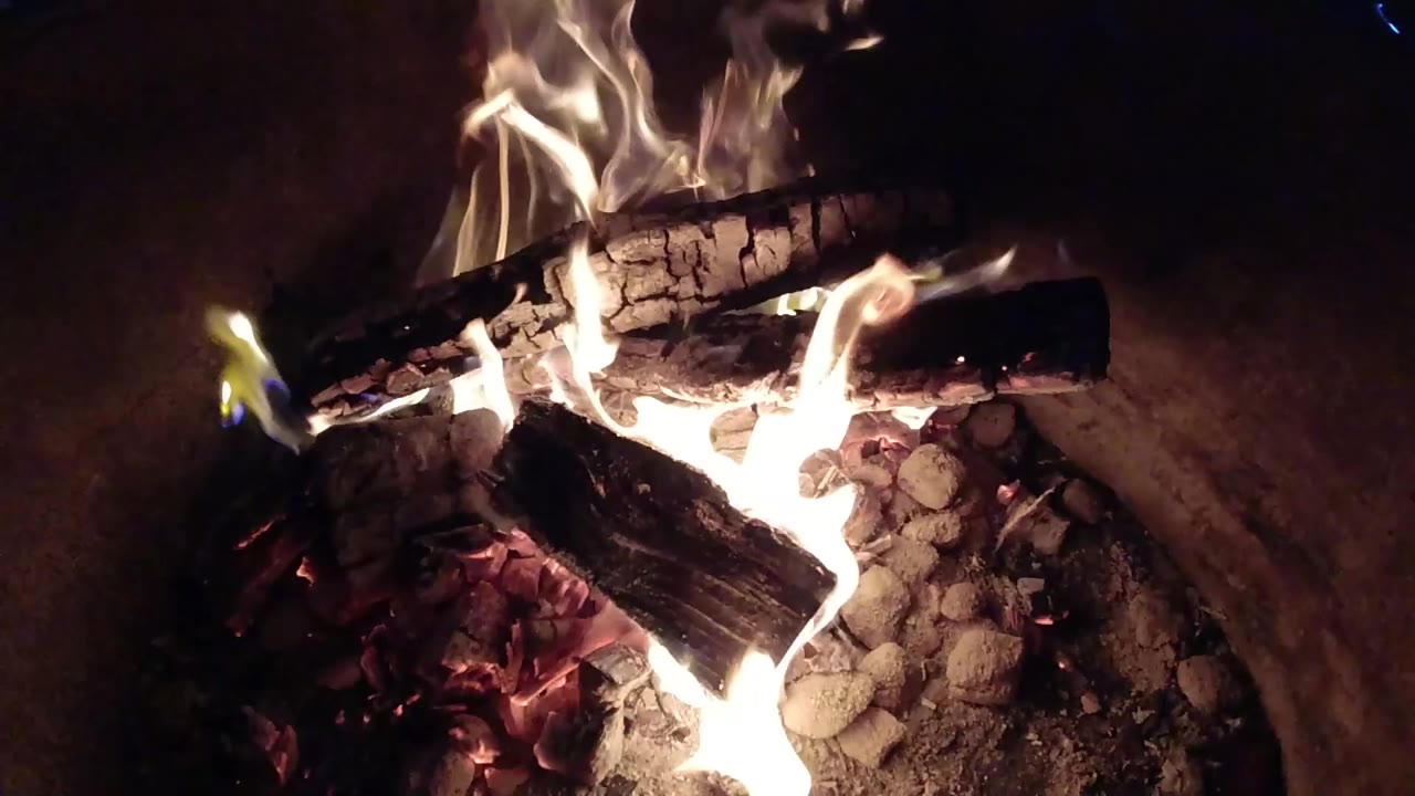 At 0:00, charred logs burn over ash and red coals in a dark fire pit in Pismo Beach, California, with no visible people.