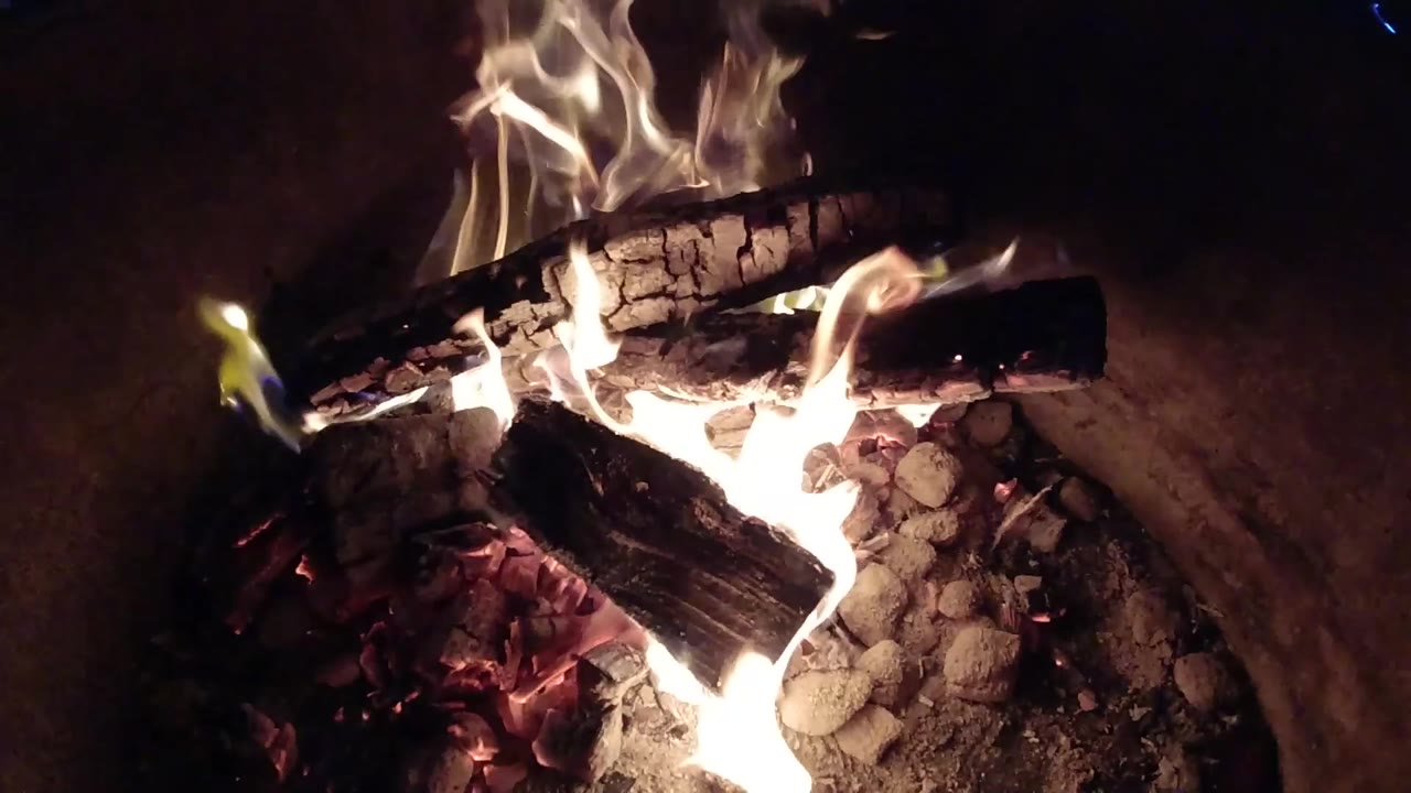 At 0:01, taller flames curl above the upper log while white ash and glowing coals remain visible below in the Pismo Beach fire pit, with no visible people.