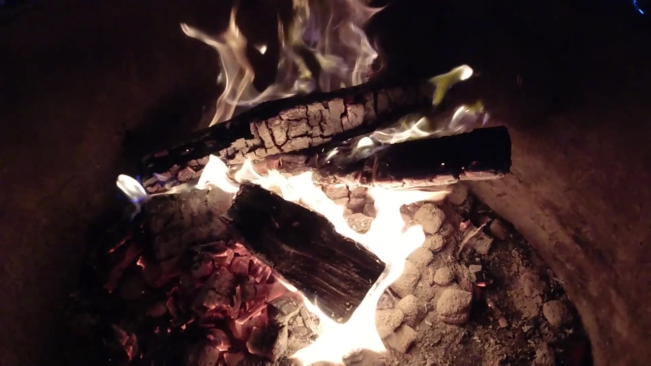 At 0:02, the flames shift to the center and right side of the stacked logs, lighting the ash and coals in the Pismo Beach fire pit, with no visible people.