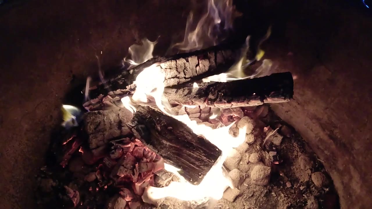 At 0:03, a bright flare appears along the left side of the upper log while the lower log and red coals remain visible in Pismo Beach, with no visible people.