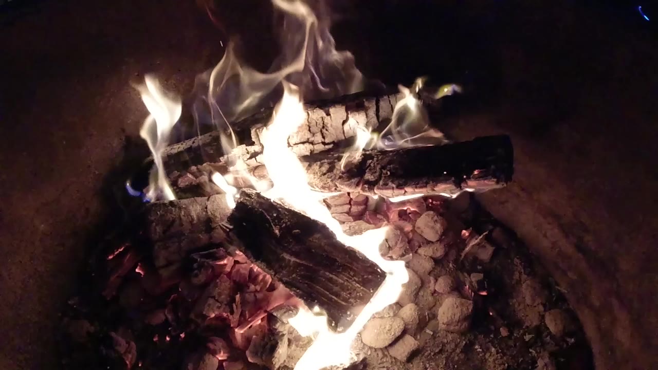Bright fire rises through stacked dark logs and scattered ash inside the fire pit, with the surrounding background mostly black and no visible people.
