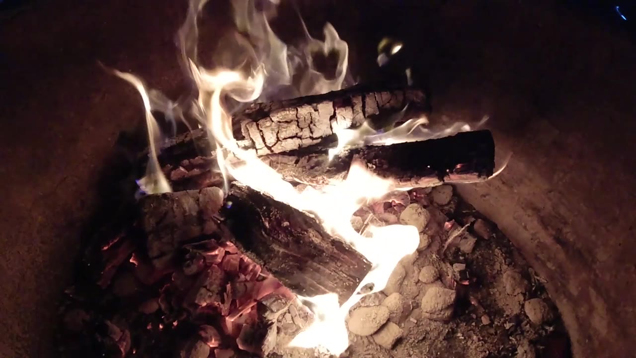 Tall flames flare below a cracked charred log, with red embers and gray ash visible near the lower edge and no visible people.
