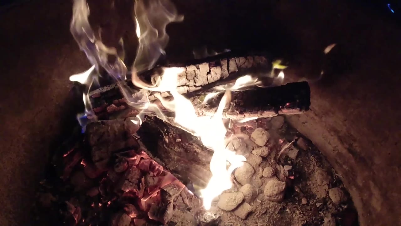 Flames curl upward from the lower edge of the fire pit toward two dark logs, with ash and ember fragments visible and no visible people.