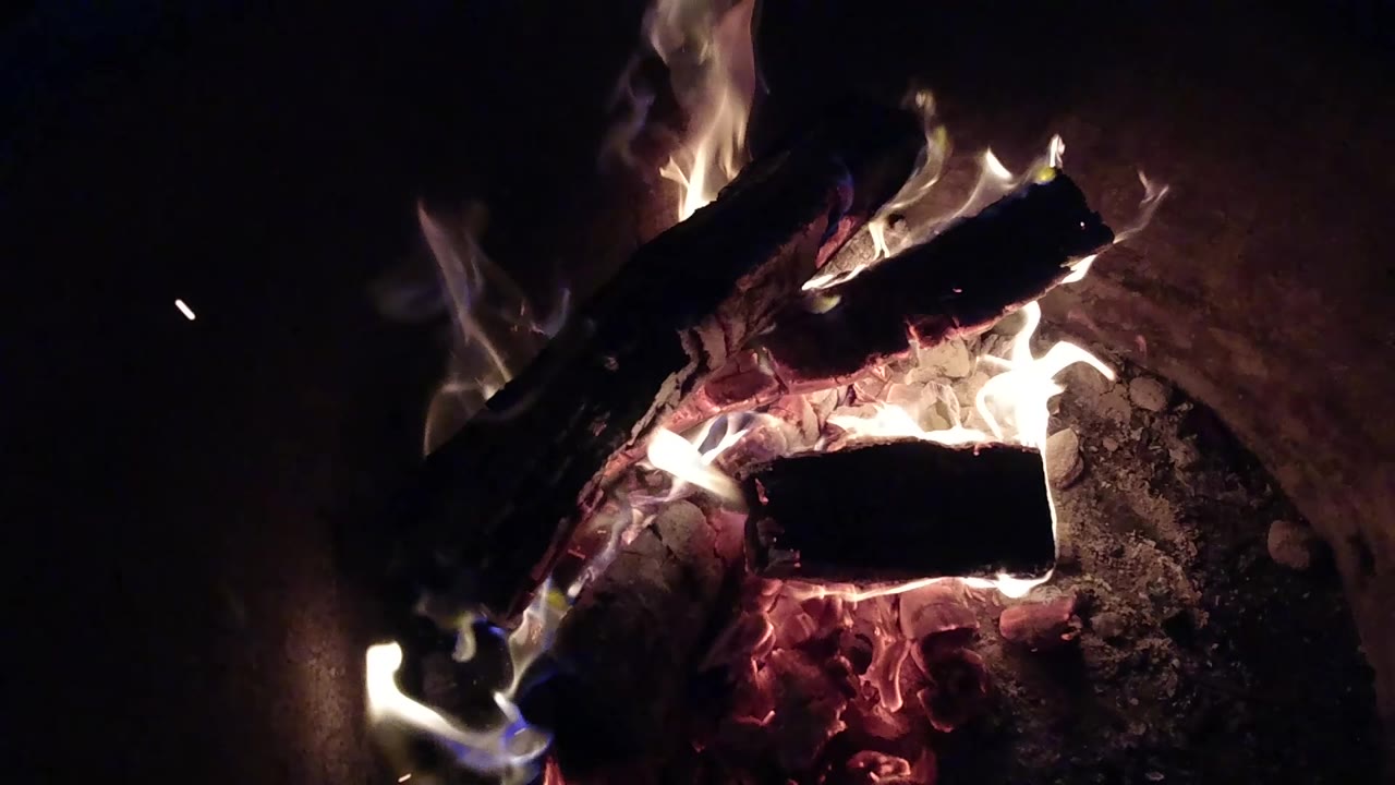 At 0:01, the fire burns brighter around the upper log in the Pismo Beach fire pit, with glowing embers below and no visible people.