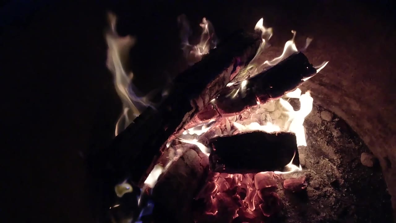 At 0:02, flames flare along the right side of the charred logs in the Pismo Beach night fire pit, with ember glow beneath and no visible people.
