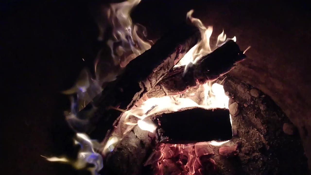 At 0:00, charred wood bars burn over red embers in Pismo Beach, California, with white flames rising from the center and no visible people.